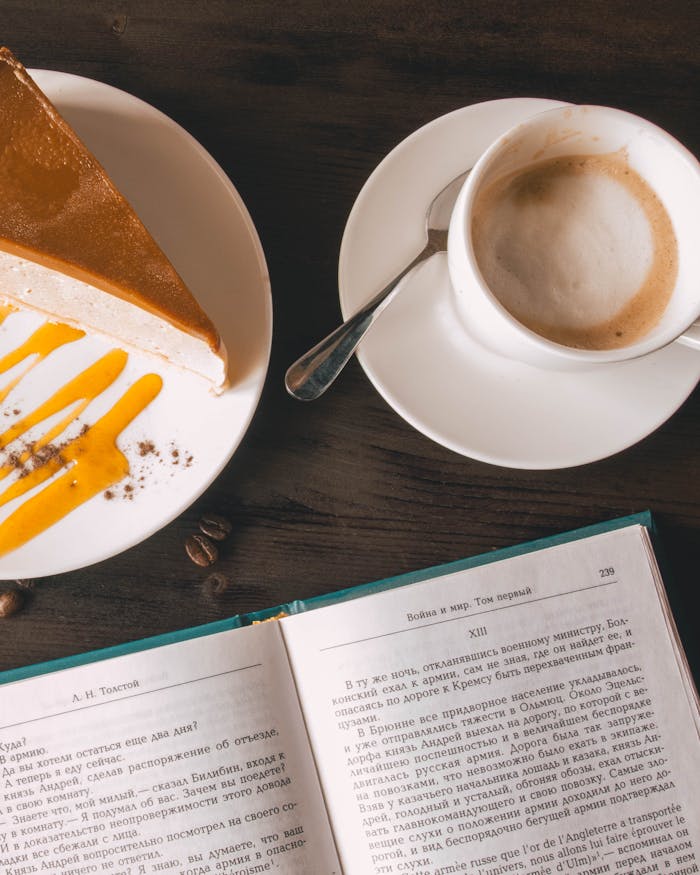 Top view of a coffee cup, cheesecake slice, and open book on a wooden table.